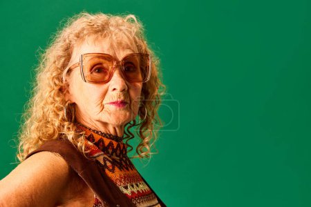Foto de Retrato de la hermosa anciana, abuela en gafas de sol con estilo y accesorios posando sobre fondo de estudio verde. Concepto de emociones humanas, moda, personas mayores, estilo de vida, creatividad. Anuncio - Imagen libre de derechos