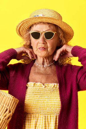 Foto de Una dama de moda. Retrato de una hermosa anciana en gafas de sol y sombrero de paja de pie sobre el fondo amarillo del estudio. Concepto de emociones humanas, moda, personas mayores, estilo de vida, creatividad. Anuncio - Imagen libre de derechos