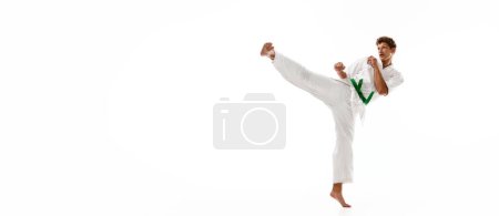 Téléchargez les photos : Jeune homme, karaté, taekwondo athlète en kimono et ceinture verte d'entraînement isolé sur fond de studio blanc. Concept d'arts martiaux, sport de combat, énergie, force, santé. Bannière. Espace de copie pour la publicité - en image libre de droit