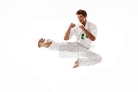Foto de Joven deportista, karateka en kimono blanco entrenando aislado sobre fondo de estudio blanco. Hombre parado en la pose de patada lateral de karate. Concepto de artes marciales, deporte de combate, energía, fuerza, salud. Anuncio - Imagen libre de derechos