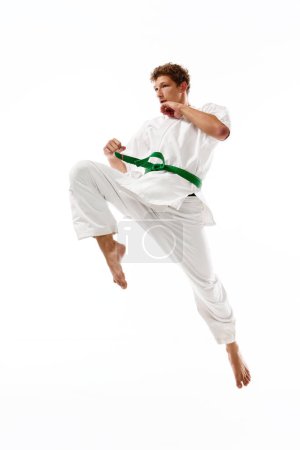 Téléchargez les photos : Jeune homme sportif, karaté, taekwondo sportif en kimono blanc en mouvement, s'entraînant isolé sur fond de studio blanc. Concept d'arts martiaux, sport de combat, énergie, force, santé. Publicité - en image libre de droit