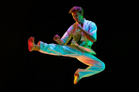 Photo for Young sportive man, karateka in white kimono training over black studio background in neon lights. Man standing in karate side kick pose. Concept of martial arts, combat sport, energy, strength. Ad - Royalty Free Image