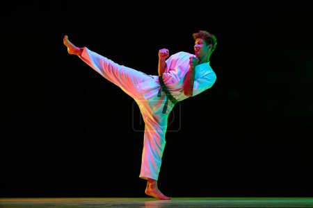 Photo for Leg kick. Professional combat sport athlete, karateka in white kimono in motion, training on black studio background in neon lights. Concept of martial arts, combat sport, energy, strength, health. Ad - Royalty Free Image