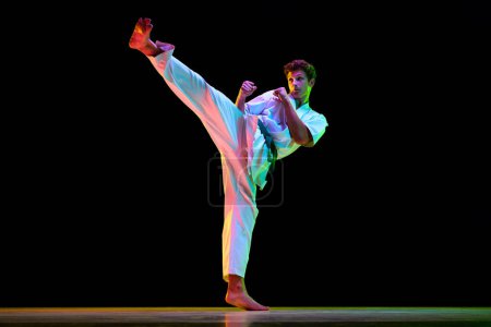 Photo for Leg kick. Professional combat sport athlete, karateka in white kimono in motion, training on black studio background in neon lights. Concept of martial arts, combat sport, energy, strength, health. Ad - Royalty Free Image