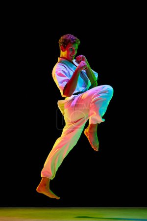 Photo for Knee kick. Young man in white kimono practicing karate punches, kicks over black studio background in neon lights. Concept of martial arts, combat sport, energy, strength, health. Ad - Royalty Free Image
