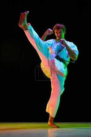 Photo for Professional combat sport. Young guy, karate athlete in motion, practicing leg kicks over black studio background in neon lights. Concept of martial arts, combat sport, energy, strength, health. Ad - Royalty Free Image