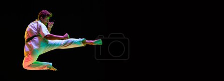 Photo for Dynamic image of young man, karate athlete in motions, training over black studio background in neon lights. Concept of martial arts, combat sport, energy, strength, health. Banner. Copy space for ad - Royalty Free Image
