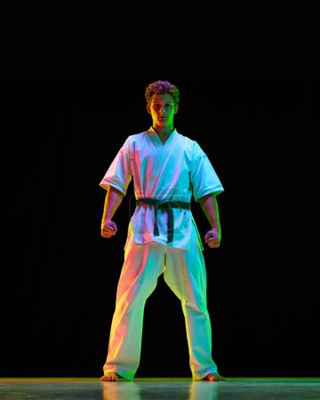 Foto de Retrato de cuerpo entero del joven deportista, atleta de karate en kimono blanco de pie sobre fondo de estudio negro con luces de neón. Concepto de artes marciales, deporte de combate, energía, fuerza, salud. Anuncio - Imagen libre de derechos