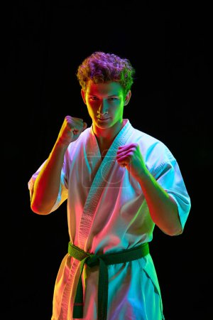 Foto de Retrato de un joven guapo en kimono blanco, karateka posando con los puños sobre el fondo negro del estudio con luces de neón. Concepto de artes marciales, deporte de combate, energía, fuerza, salud. Anuncio - Imagen libre de derechos