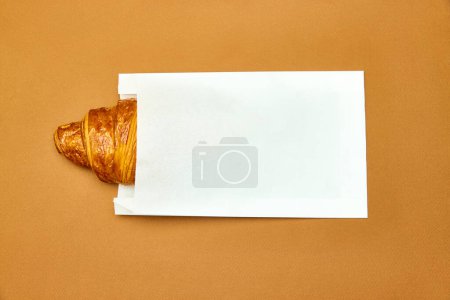 Photo for Piece of crispy, freshly baked croissant peaking out paper packaging isolated over brown background. Take away. Concept of food, bakery, breakfast ideas, taste, freshness. Poser. Copy space for ad - Royalty Free Image