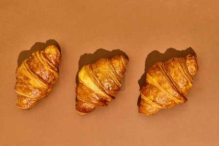 Téléchargez les photos : Trois croissants croustillants fraîchement cuits isolés sur fond brun. Goût délicieux, salé et sucré. Concept de nourriture, boulangerie, idées de petit déjeuner, goût, fraîcheur. Poser. Espace de copie pour la publicité - en image libre de droit
