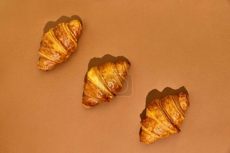 Photo for Three crispy freshly baked croissants isolated over brown background. Yummy taste, salted and sweet. Concept of food, bakery, breakfast ideas, taste, freshness. Poser. Copy space for ad - Royalty Free Image