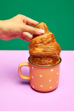 Photo for Woman dipping fresh croissant into cup with coffee, latte isolated over pink green background. Concept of food, bakery, breakfast ideas, taste, freshness. Poser. Copy space for ad - Royalty Free Image