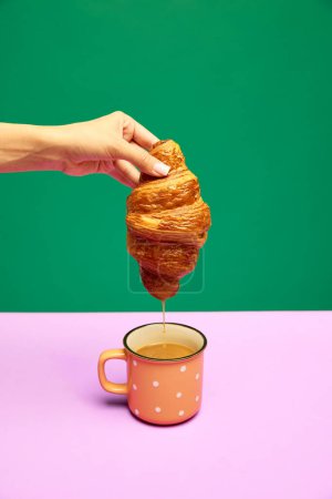 Photo for Woman dipping fresh croissant into cup with coffee, latte isolated over pink green background. Concept of food, bakery, breakfast ideas, taste, freshness. Poser. Copy space for ad - Royalty Free Image