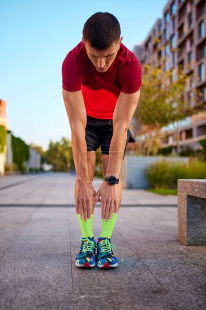 Photo for Warming-up before training. Man in sportswear standing on empty street, doing toe touching exercises before running. Concept of sport, active and healthy lifestyle, competition, dynamics, marathon - Royalty Free Image