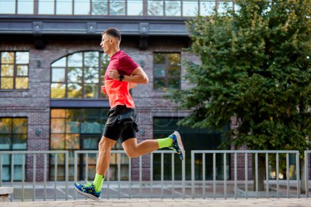 Photo for Athletic young man, runner in motion training outdoors in city on warm morning. Running along empty street. Concept of sport, active and healthy lifestyle, competition, dynamics, marathon - Royalty Free Image