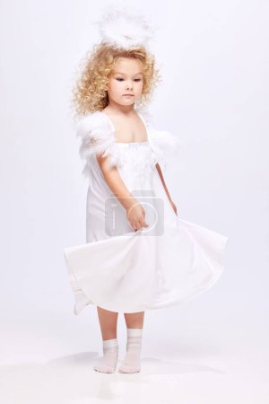 Téléchargez les photos : Belle, mignonne petite fille enfant en robe blanche à l'image d'ange debout isolé sur fond de studio blanc. Concept d'enfance, imagination, fantaisie, mode et beauté, vacances - en image libre de droit