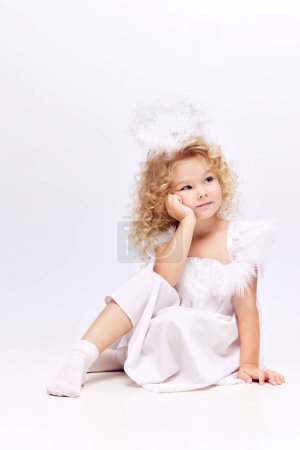 Foto de Linda, adorable niña, niño en vestido blanco y halo ángeles sentado con la cara de ensueño aislado en el fondo del estudio blanco. Concepto de infancia, imaginación, fantasía, moda y belleza, vacaciones - Imagen libre de derechos