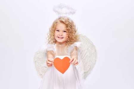 Téléchargez les photos : Joyeuse Saint-Valentin. Petit bébé Cupidon, belle fille en robe blanche tenant coeur en papier isolé sur fond de studio blanc. Concept d'enfance, imagination, fantaisie, bonheur, vacances - en image libre de droit