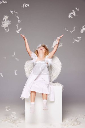 Téléchargez les photos : Joyeux, souriant, petite fille positive, enfant en costume d'ange assis isolé sur fond de studio gris avec des plumes volantes. Concept d'enfance, imagination, fantaisie, mode, vacances - en image libre de droit