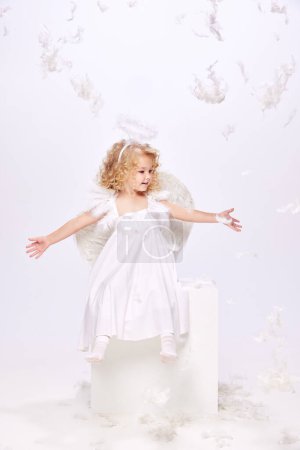 Téléchargez les photos : Joyeux, souriant, petite fille positive, enfant en costume d'ange assis isolé sur fond de studio blanc avec des plumes volantes. Concept d'enfance, imagination, fantaisie, vacances, vacances - en image libre de droit