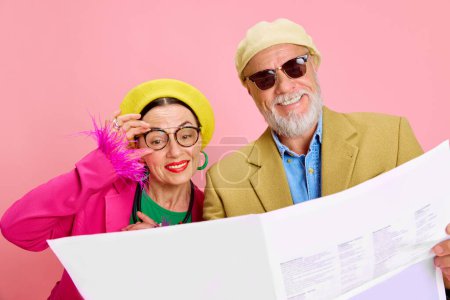 Téléchargez les photos : Nouvelles excitées. Joyeux couple de personnes âgées souriant dans des accessoires élégants, béret et lunettes de soleil, lecture de journal sur fond de studio rose. Concept de beauté et de mode, relation, style moderne, âge - en image libre de droit