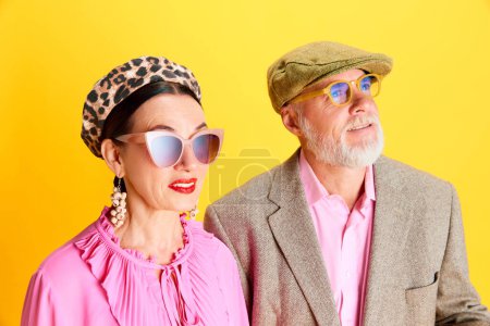 Téléchargez les photos : Belle femme âgée en béret, lunettes de soleil et chemisier rose debout avec un bel homme barbu avec casquette et veste sur fond jaune. Concept de beauté, mode, relation, style moderne, âge - en image libre de droit