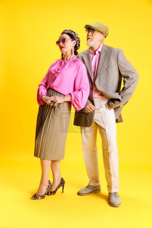Photo for Family. Stylish, elegant grandparents, man and woman in matching clothes and accessories standing against yellow studio background. Concept of beauty and fashion, relationship, modern style, age - Royalty Free Image
