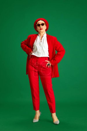 Foto de De larga duración. Mujer elegante, hermosa y madura de unos 60 años, con un elegante traje rojo, boina y gafas de sol, de pie sobre un fondo verde del estudio. Concepto de belleza y moda, estilo moderno, edad - Imagen libre de derechos