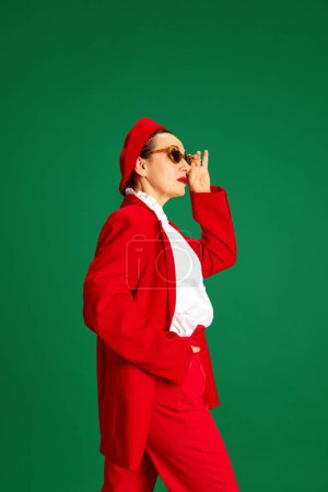 Téléchargez les photos : Vue de côté de la femme élégante dans les années 60, portant élégant, costume rouge vif et béret, posant sur fond de studio vert. Concept de beauté et de mode, style moderne, âge. Femme d'affaires - en image libre de droit