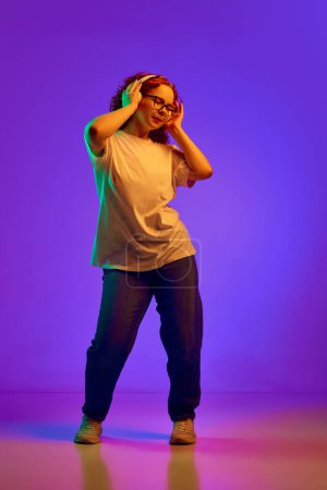 Photo for Full-length. Teen girl with curly hair listening to music in headphones and dancing against purple background in neon light. Concept of human emotions, lifestyle, youth, facial expression. Ad - Royalty Free Image