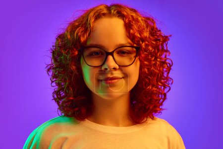 Photo for Portrait of smiling, cheerful teen girl with curly redhead hair, in glasses, lookin at camera on purple background in neon light. Concept of human emotions, lifestyle, youth, facial expression. Ad - Royalty Free Image