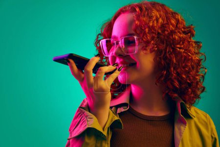 Photo for Smiling teen girl with curly hair recording voice message on mobile phone over cyan background in neon light. Online communication. Concept of human emotions, lifestyle, youth, facial expression. Ad - Royalty Free Image