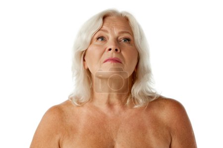Téléchargez les photos : Belle femme âgée dans la soixantaine debout avec les épaules nues, regardant vers le haut sur fond de studio blanc. Concept de beauté naturelle, processus de vieillissement, beauté âgée, cosmétologie, soins de la peau - en image libre de droit