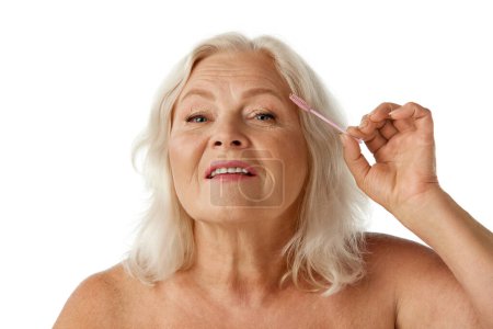 Photo for Elegant, beautiful senior woman taking care after brows, brushing with brush against white studio background. Concept of natural beauty, aging process, elderly beauty, cosmetology, skincare - Royalty Free Image