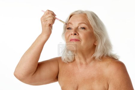Téléchargez les photos : Elégante, belle femme âgée se maquillant nue, appliquant des ombres à paupières sur fond de studio blanc. Concept de beauté naturelle, processus de vieillissement, beauté âgée, cosmétologie, soins de la peau - en image libre de droit