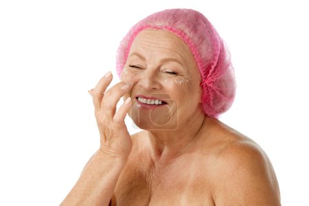 Téléchargez les photos : Souriante, belle femme âgée prenant soin de la peau après, appliquer sous la crème pour les yeux sur fond blanc studio. Concept de beauté naturelle, processus de vieillissement, beauté âgée, cosmétologie, soins de la peau - en image libre de droit