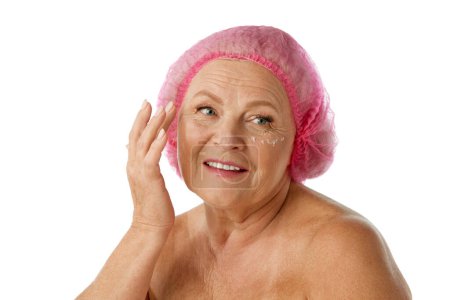 Téléchargez les photos : Souriante, belle femme âgée prenant soin de la peau après, appliquer sous la crème pour les yeux sur fond blanc studio. Concept de beauté naturelle, processus de vieillissement, beauté âgée, cosmétologie, soins de la peau - en image libre de droit