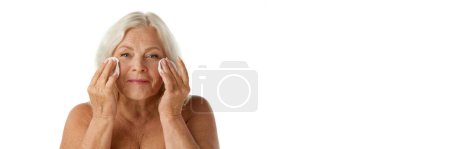 Téléchargez les photos : Belle femme âgée avec des rides, se démaquiller avec des tampons de coton sur fond de studio blanc. Concept de beauté naturelle, processus de vieillissement, beauté âgée, cosmétologie, soins de la peau. Bannière - en image libre de droit
