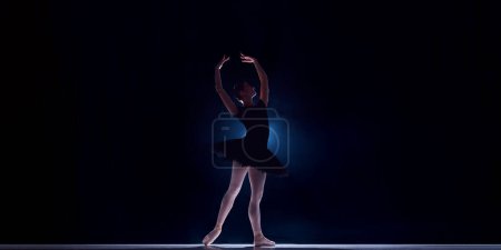 Téléchargez les photos : Belle et élégante jeune femme, danseuse de ballet professionnelle en tutu dansant sur scène sur fond sombre. Concept de danse classique, art et grâce, beauté, chorégraphie, inspiration - en image libre de droit