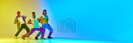 Photo for Hip hop dancers. Three men in stylish vintage sportswear and accessories posing against gradient yellow blue background in neon light. Concept of sportive and active lifestyle, humor, retro style. Ad - Royalty Free Image