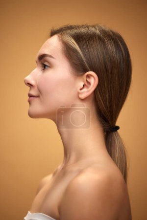 Photo for Profile view of tender, attractive young girl with ponytail, standing with bare shoulders isolated over light brown background. Concept of natural beauty, skincare, cosmetics and cosmetology - Royalty Free Image