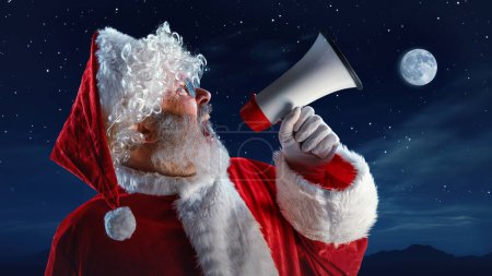 Téléchargez les photos : Homme aîné, le Père Noël crie émotionnellement en mégaphone sur le ciel étoilé avec la lune. C'est l'heure des miracles. Concept de saison d'hiver, vacances, fantaisie, joie et plaisir, Noël - en image libre de droit