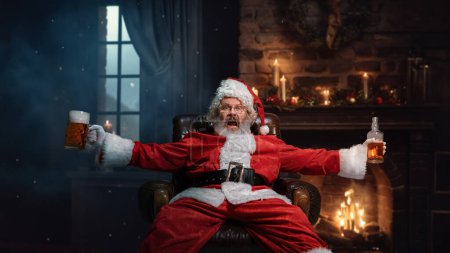 Foto de Hombre mayor emocional, Santa Claus sentado cerca de la chimenea con taza de cerveza y botella de coñac en la noche. Es hora de celebrar. Concepto de temporada de invierno, vacaciones, fantasía, alegría y diversión, Navidad - Imagen libre de derechos