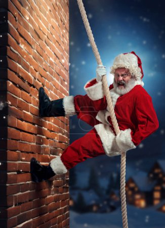 Photo for Senior bearded man, Santa Claus climbing upwards the brick wall at night. Entering house through chimney. Concept of winter season, holidays, fantasy, joy and fun, Christmas - Royalty Free Image