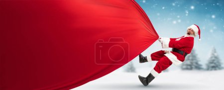 Photo for Emotional senior man in costume, Santa Claus carrying giant bag with preset on snowy forest. Big sales, shopping. Concept of winter season, holidays, fantasy, joy and fun, Christmas - Royalty Free Image