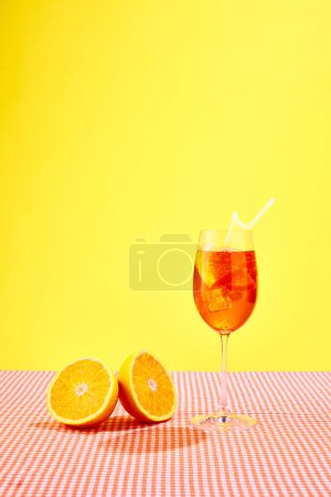 Foto de Copa de cóctel Aperol spritz con naranjas de pie sobre mantel a cuadros sobre fondo amarillo. Vibra de verano. Concepto de bebidas alcohólicas, fiesta, vacaciones, bar, mezcla. Cartel. Copiar espacio para anuncio - Imagen libre de derechos