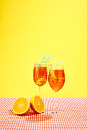 Téléchargez les photos : Deux verres d'Aperol spritz cocktail avec des oranges debout sur la nappe à carreaux sur fond jaune. Ambiance estivale. Concept de boissons alcoolisées, fête, vacances, bar, mix. Affiche. Espace de copie pour la publicité - en image libre de droit
