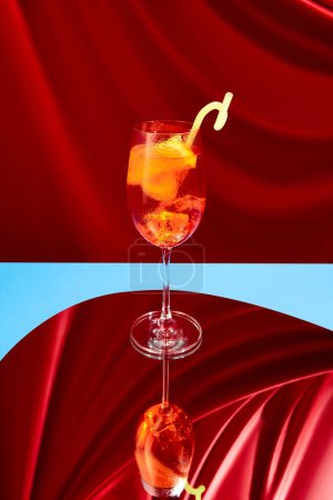 Téléchargez les photos : Délicieux, frais, rafraîchissant cocktail Aperol Spritz debout sur miroir sur fond de soie rouge. Élégance. Concept de boissons alcoolisées, fête, vacances, bar, mix. Affiche. Espace de copie pour la publicité - en image libre de droit