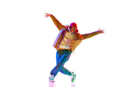 Téléchargez les photos : Sportif, élégant jeune homme en tenue de sport danse de rue danse isolé sur fond de studio blanc dans la lumière au néon. Concept de danse contemporaine, style de rue, mode, passe-temps, jeunesse. Publicité - en image libre de droit